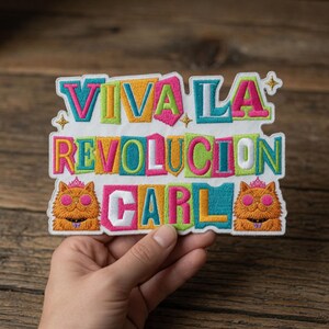 Könnte beinhalten: Gestickter Aufnäher mit dem Text "VIVA LA REVOLUCION CARL" in bunten Blockbuchstaben. Der Aufnäher zeigt zwei Cartoon-Katzen-Illustrationen mit Sonnenbrillen und Kronen. Der Aufnäher ist rechteckig mit weißem Rand.