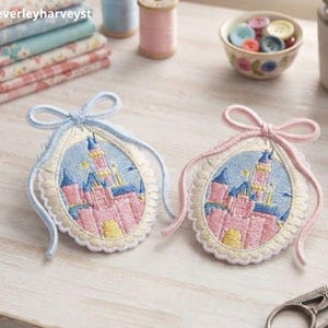 Peut inclure: Deux écussons brodés ovales représentant un château rose et bleu. L'un est entouré d'un cordon bleu clair avec un nœud, l'autre d'un cordon rose clair avec un nœud. Les deux écussons présentent des bords festonnés et des détails brodés.