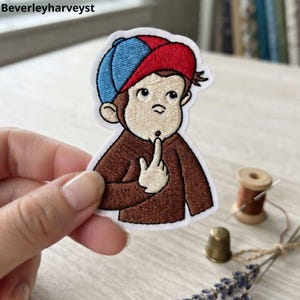 May include: An embroidered patch featuring a cartoon monkey with light brown fur, wearing a red and blue cap. The monkey has a cream-coloured face and chest, with one finger raised to its chin in a thoughtful expression. The patch has a white border.