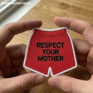 Può includere: Una toppa rossa a forma di pantaloncino da boxe con bordatura bianca. La toppa presenta le parole "RESPECT YOUR MOTHER" in nero, ricamate sul davanti. La toppa è tenuta da due mani.