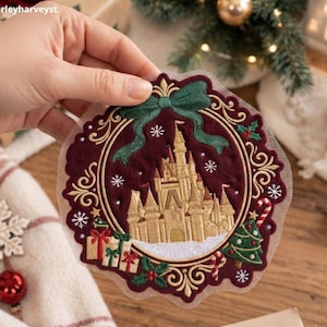 Puede incluir: Un parche bordado circular que presenta un castillo dorado sobre un fondo burdeos intenso. El diseño incluye un lazo verde en la parte superior, un árbol de Navidad decorado, regalos, acebo y copos de nieve blancos, enmarcado por un borde dorado.