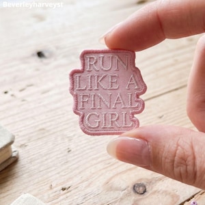 May include: A light pink embroidered patch with a rectangular shape and rounded corners. The patch features the white stitched text "RUN LIKE A FINAL GIRL" in a bold, sans-serif font. The edges of the patch are finished with a matching pink border.
