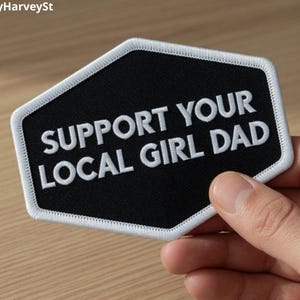 May include: Black and white embroidered patch with the text "SUPPORT YOUR LOCAL GIRL DAD". The patch has a black background with white lettering and a white border. The patch is held in a hand.