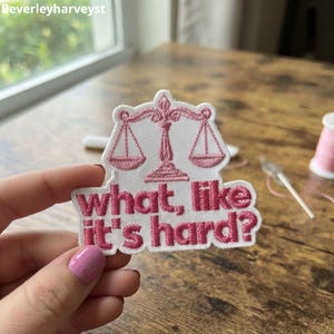 以下が含まれることがあります： 白い刺繍ワッペン。ピンク色の天秤のアイコンと、その下にピンク色の刺繍文字で「what, like it's hard?」と記されている。ワッペンはデザインの輪郭に沿ったダイカット形状で、白い縁取りが施されている。