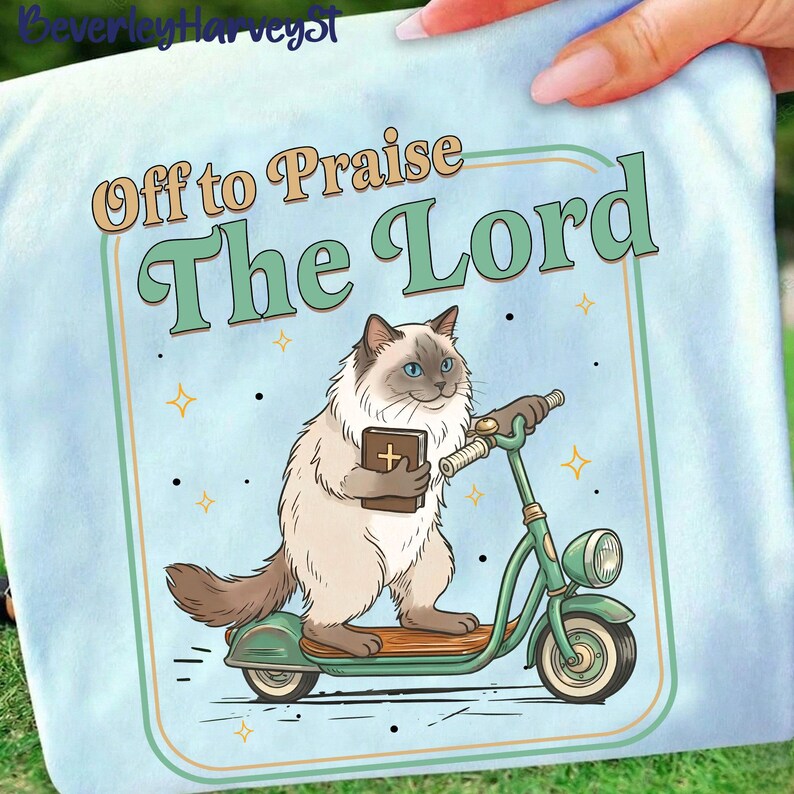 May include: Light blue t-shirt with a cartoon cat riding a green scooter and holding a book. The text "Off to Praise The Lord" is above the cat. The design includes stars and a light green border.