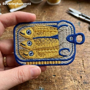 May include: A rectangular embroidered patch featuring three yellow fish with blue eyes inside a silver-colored tin can. The design is outlined in a bold blue thread and includes a blue pull-tab on the right side. The patch has a textured finish.