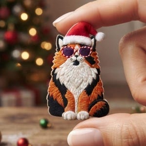 May include: An embroidered patch of a calico cat with black, orange, and white fur. The cat wears a red and white Santa hat and purple-tinted sunglasses with a red and white striped frame. The patch features detailed stitching throughout.