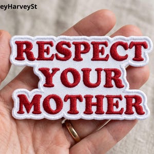 May include: A white patch with red embroidered text that reads "RESPECT YOUR MOTHER". The patch has a white border and is held in a hand.