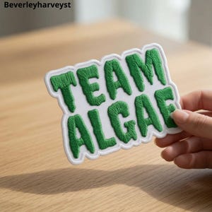 May include: A white embroidered patch with a scalloped edge featuring the text TEAM ALGAE in bold, textured green thread. The letters have a slightly irregular, organic shape, and the patch is designed to be applied to fabric surfaces.