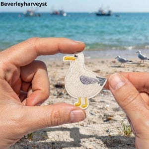 May include: An embroidered patch shaped like a seagull with a cream-coloured body, a yellow beak and feet, and a grey wing. The patch features a simple stitched design and is held up against a blurred coastal background with the text Beverleyharveyst.