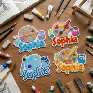 May include: Four embroidered patches featuring the name Sophia in various colors, each depicting a paint palette, a paintbrush, and a paint jar. The patches are arranged on a wooden surface surrounded by art supplies including pencils and paint tubes.