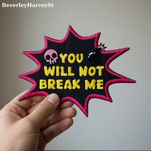 May include: A black patch with a pink and yellow design. The patch features a pink skull, a black bomb, and the yellow text "YOU WILL NOT BREAK ME". The patch is in the shape of a starburst.