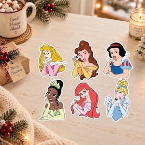 Op de afbeelding: Een set van zes gestanste vinylstickers met geïllustreerde personages: Aurora met blond haar, Belle met bruin haar, Sneeuwwitje met zwart haar, Tiana met een groene tiara, Ariël met rood haar en Assepoester met een blond opgestoken kapsel.