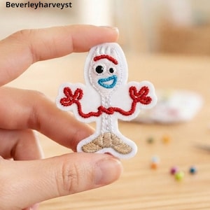 May include: An embroidered patch featuring a white spork character with a red pipe cleaner arm, one large black eye, one small black eye, and a blue mouth. The character has tan feet and is outlined in white thread on a white fabric backing.