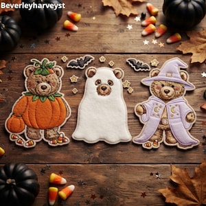 May include: Three embroidered patches featuring brown teddy bears in Halloween costumes: one dressed as a pumpkin, one as a ghost, and one as a wizard in a purple robe. The patches are arranged on a wooden surface with scattered candy corn.