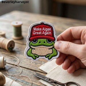 May include: An embroidered patch featuring a green frog wearing a red baseball cap with the white text Make Algae Great Again. The patch has a white border and depicts the frog with a light tan belly and dark eyes, designed for sewing or ironing.