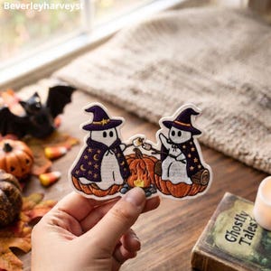 May include: An embroidered patch featuring two white ghosts wearing purple witch hats and capes decorated with gold stars and moons. The ghosts sit on orange pumpkins, roasting marshmallows over a small campfire between them on a white background.