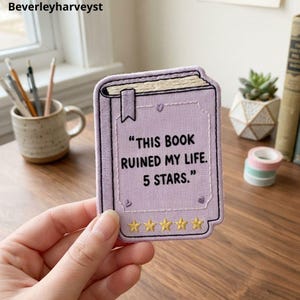 Peut inclure: Un écusson brodé rectangulaire en forme de livre avec une couverture violet clair. Le texte indique : "THIS BOOK RUINED MY LIFE. 5 STARS." Il comporte un petit marque-page, trois minuscules cœurs brodés et cinq étoiles jaunes en bas.