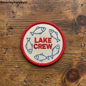 Op de afbeelding: Een ronde geborduurde patch met een crèmekleurige achtergrond en een rode rand. De tekst "LAKE CREW" is in het midden in het rood geborduurd, omringd door vier blauwe geborduurde visomtrekken in een cirkelvormig patroon.