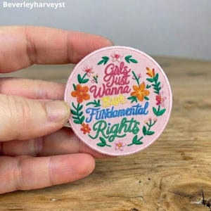 Könnte beinhalten: Ein runder bestickter Aufnäher auf hellrosa Stoffhintergrund. Der Text Girls Just Wanna Have Fundamental Rights ist in rosa, gelbem und blauem Garn gestickt. Das Design ist von bunten gestickten Blumen und grünen Blättern umgeben.