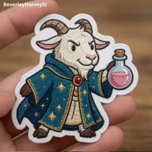 Op de afbeelding: Een geborduurde patch met een witte geit in een donkergroenblauw gewaad met gouden sterpatronen en een rode edelsteensluiting. De geit houdt een klein glazen flesje met roze vloeistof vast. De patch heeft een witte rand en achtergrond.