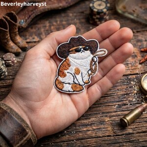Puede incluir: Un parche bordado que muestra un gato blanco y naranja con un sombrero de vaquero marrón y gafas de sol negras, sosteniendo un lazo. El parche tiene un borde blanco y tiene la forma de la figura del gato. El texto Beverleyharveyst es visible.