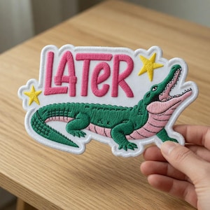 May include: Embroidered patch featuring a green alligator with a pink underbelly and open mouth. The word "LATER" is in pink, with yellow stars on a white background. The patch is held by a hand.