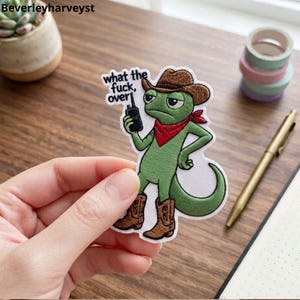 May include: An embroidered patch featuring a green chameleon wearing a brown cowboy hat, brown cowboy boots, and a red bandana. The chameleon holds a black walkie-talkie and the text "what the fuck, over" is embroidered in black above the device.