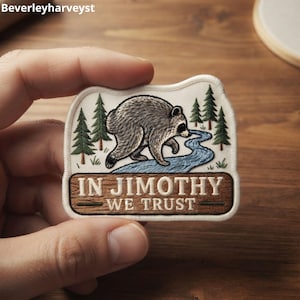 May include: A rectangular embroidered patch with a cream-colored background featuring a grey raccoon walking near a blue river and green pine trees. Below the scene, a brown wooden-textured banner displays the white text IN JIMOTHY WE TRUST.