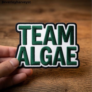 May include: A rectangular embroidered patch featuring the text TEAM ALGAE in dark green block letters on a white background, outlined with a thick black border. The patch has a textured surface and is designed for attachment to fabric items.