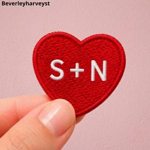May include: A red heart-shaped embroidered patch featuring the white stitched letters S + N in the centre. The patch has a textured fabric surface with a defined border and is presented against a solid light pink background with the text Beverleyharveyst.