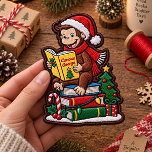 Puede incluir: Un parche bordado que muestra a Curious George con un gorro y bufanda de Papá Noel rojos, leyendo un libro amarillo titulado Curious George. El mono está sentado sobre tres libros apilados junto a un árbol de Navidad, un bastón de caramelo y regalos.