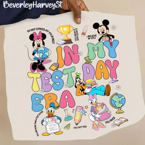 Peut inclure: Un t-shirt beige avec l'inscription colorée "IN MY TEST DAY ERA". Le design présente des personnages de dessins animés comme Minnie, Mickey, Dingo, Donald et Daisy, ainsi que des illustrations scolaires, comme un globe et des livres.