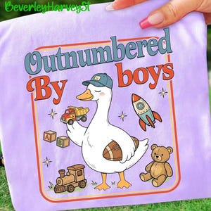 May include: A lilac t-shirt with a cartoon goose wearing a cap and holding a toy truck and a football. The text "Outnumbered By Boys" is displayed in a retro font, along with a rocket, a teddy bear, a train, and alphabet blocks.