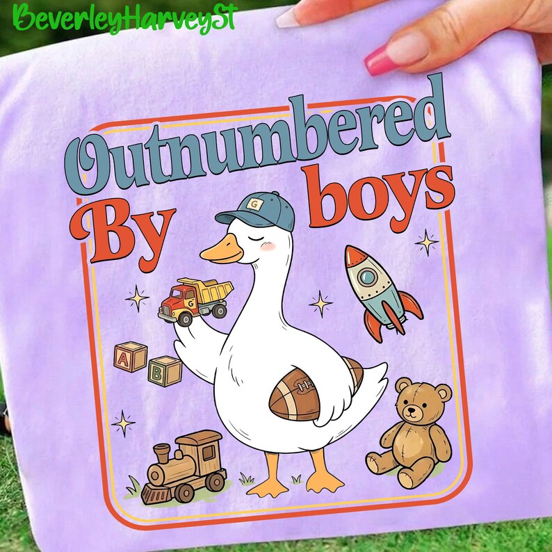 May include: A lilac t-shirt with a cartoon goose wearing a cap and holding a toy truck and a football. The text "Outnumbered By Boys" is displayed in a retro font, along with a rocket, a teddy bear, a train, and alphabet blocks.