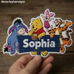 Puede incluir: Un parche bordado que muestra a Igor, Winnie the Pooh con un tarro de miel, Piglet y Tigger rodeando un letrero de madera azul oscuro con el texto blanco Sophia. El parche tiene un borde blanco y se sostiene sobre un fondo de madera.