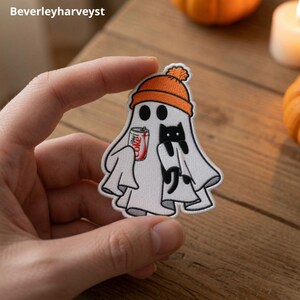 May include: An embroidered patch featuring a white ghost wearing an orange beanie. The ghost holds a small silver soda can with red text and embraces a small black cat. The design has a white border and is shaped like a classic sheet ghost.