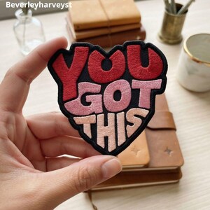 May include: A heart-shaped embroidered patch with a black border and the text YOU GOT THIS. The word YOU is in red, GOT is in pink, and THIS is in light peach. The embroidery features a dense, textured stitch pattern throughout the letters.