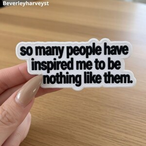 May include: A white embroidered patch with a black stitched border and black text that reads: so many people have inspired me to be nothing like them. The patch has a die-cut shape that follows the contour of the three lines of text.