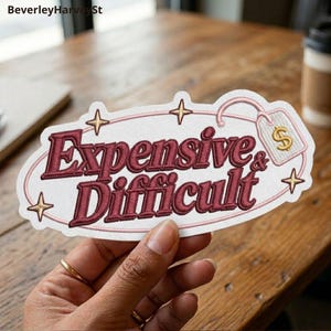 May include: An embroidered patch with a white background and a pink oval border. The text reads Expensive & Difficult in dark red, bold, serif lettering. Four gold stars surround the text, and a pink price tag with a gold dollar sign is attached.