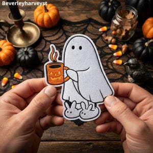 Op de afbeelding: Een geborduurde patch met een wit spookje met zwarte ogen, witte konijnenpantoffels aan en een oranje mok met een pompoengezicht waar stoom uit komt. De patch is afgewerkt met een zwarte rand en witte stiksels.