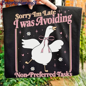 May include: Black t-shirt with a cartoon goose wearing a pink bow. The text reads "Sorry I'm Late I Was Avoiding Non-Preferred Tasks" in pink and beige. The design includes stars, hearts, and flowers.