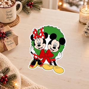 May include: A die-cut sticker featuring Mickey and Minnie Mouse standing together inside a green Christmas wreath. Minnie wears her signature red and white polka dot bow and dress, while Mickey wears his yellow shoes. A large red bow is at the base.
