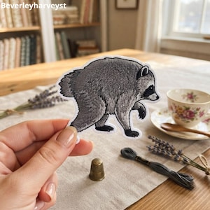 May include: A die-cut sticker featuring a detailed illustration of a gray raccoon walking. The raccoon has a black mask around its eyes, black paws, and a fluffy tail. The artwork uses fine lines to depict the texture of the animal's fur.