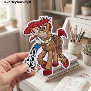 Peut inclure: Un écusson brodé représentant un personnage de dessin animé aux cheveux roux et au chapeau rouge serrant un cheval brun dans ses bras. Le personnage porte une chemise blanche, un gilet bleu et un pantalon blanc à taches de vache noires.