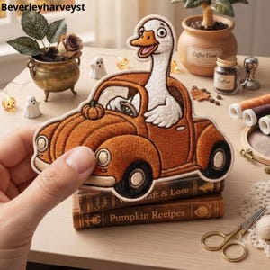 Puede incluir: Un parche bordado que muestra a un ganso blanco conduciendo un coche naranja con forma de calabaza. El ganso tiene el pico abierto y sujeta un volante. El parche se sostiene frente a un fondo de libros, una planta pequeña y artículos de costura.
