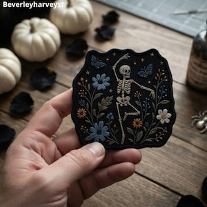 May include: A black fabric patch featuring an embroidered design of a dancing skeleton surrounded by blue and orange flowers, green leaves, and two blue butterflies. The patch has a scalloped edge and includes small embroidered star accents.