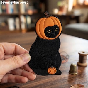 Pode incluir: Um emblema bordado apresentando um gato preto com uma cabeça de abóbora laranja brilhante e uma pequena abóbora laranja aos seus pés. O emblema tem uma borda preta e é segurado contra um fundo desfocado. O texto Beverleyharveyst é visível.