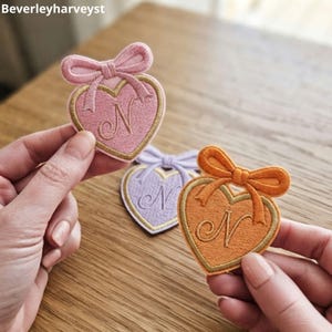 以下が含まれることがあります： 上部にリボン、中央に筆記体のNの文字が刺繍された、ハート型のワッペン3枚。ピンク、薄紫、オレンジの3色で、それぞれ金色の縁取りと細かな刺繍が施されています。手芸用の装飾として使用できる、布製のハート型ワッペンです。
