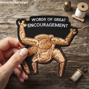 Puede incluir: Un parche bordado negro que muestra una criatura de color tostado con textura agrietada y múltiples extremidades levantadas. El texto "WORDS OF GREAT ENCOURAGEMENT" está bordado en letras mayúsculas blancas sobre el fondo negro superior.